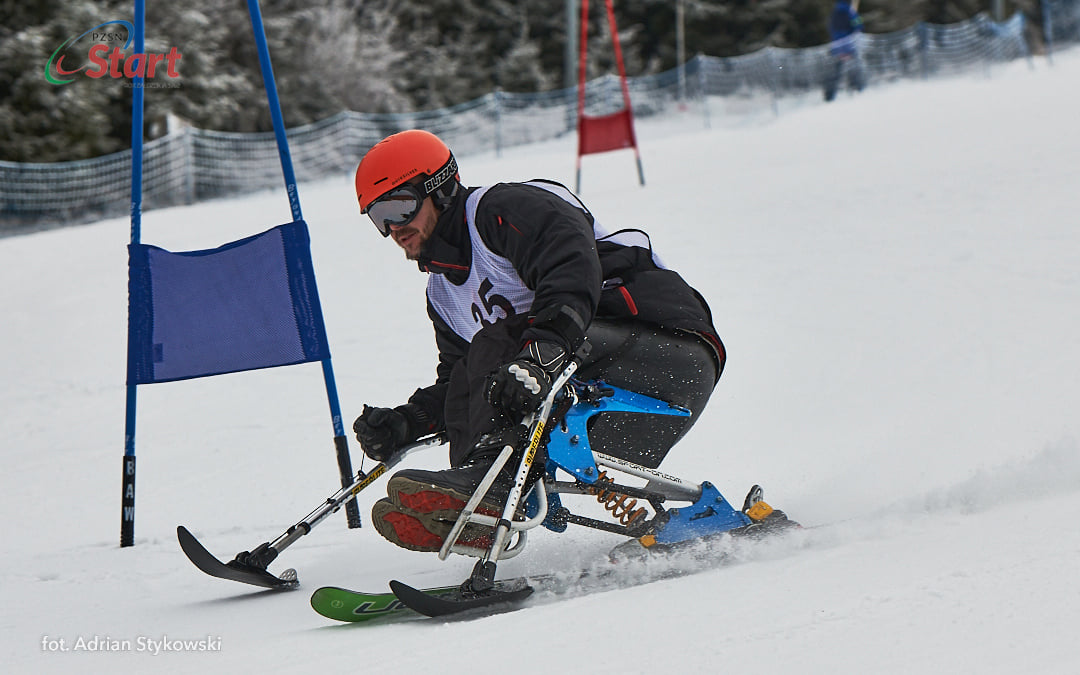 Zdjęcie przedstawia mężczyznę na siedzisku z nartą zjazdową w trakcie zawodów.