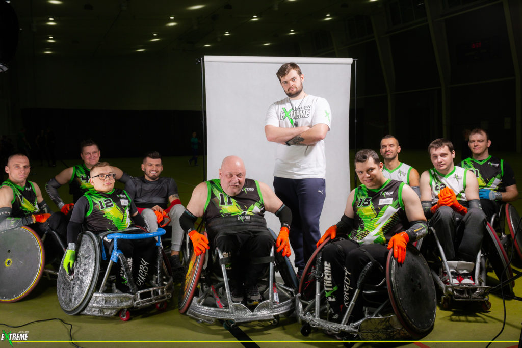 Na zdjęciu znajduje się męska drużyna rugby. Zawodnicy znajdują się na specjalistycznych wózkach i pozują do zdjęcia wraz z trenerem.