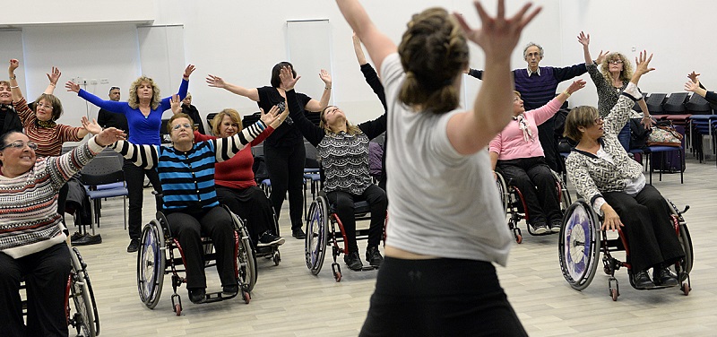 Na zdjęciu znajduje się grupa tancerzy na wózkach w trakcie ćwiczenia choreografii. Wszyscy mają ręce uniesione w górę. 