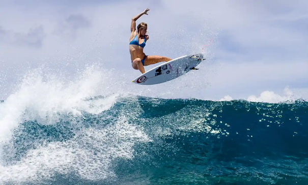 Zdjęcie przedstawia kobietę na desce surfingowej, która surfuje na dużej fali. 