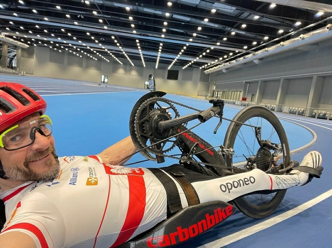 Na zdjęciu znajduje się uśmiechnięty mężczyzna (Rafał Wilk) na rowerze ręcznym, przebywa na hali sportowej.