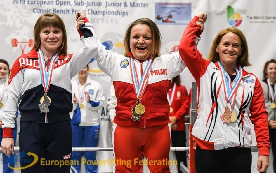 Na zdjęciu znajdują się trzy kobiety z medalami na szyjach, które trzymają się za ręce i unoszę je razem w górę. W środku znajduje się Justyna Kozdryk.