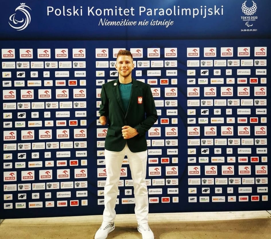 Na zdjęciu znajduje się Bartłomiej Mróz, który stoi na tle ścianki promocyjnej Polskiego Komitetu Paraolimpijskiego.