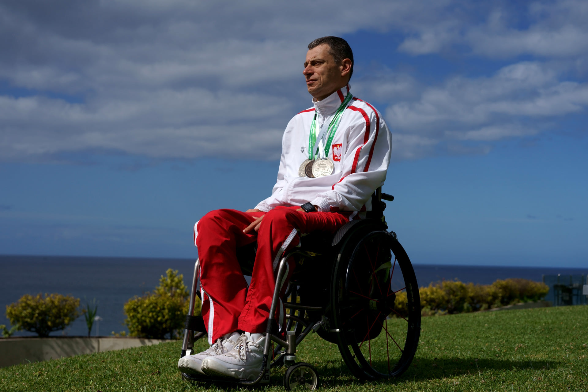 Zdjęcie przedstawia Jacka Czecha w sportowym stroju i z medalami na szyi, który znajduje się na wózku. Jest sfotografowany na zielonej trawie, na tle jasnoniebieskiego nieba.