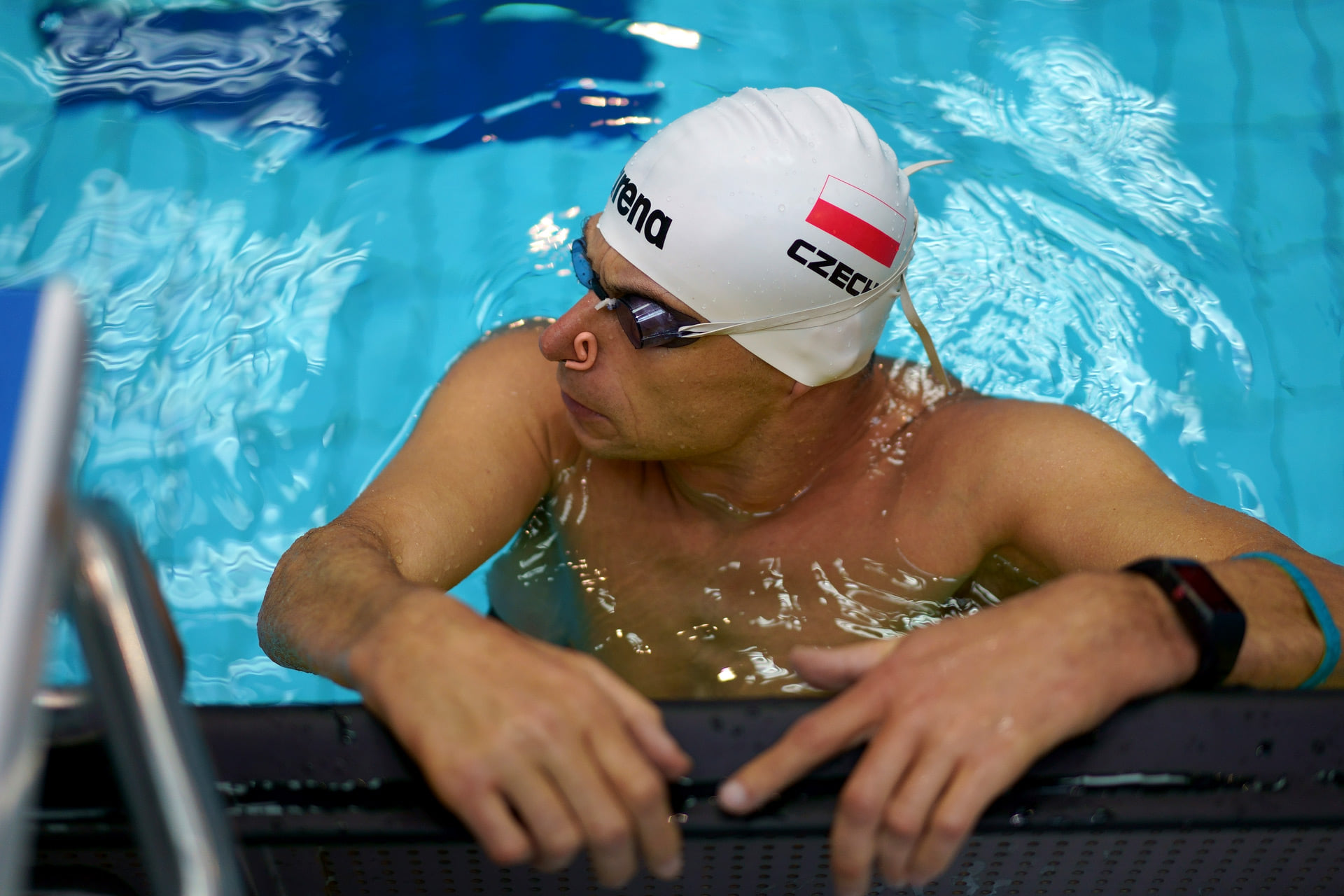 Na zdjęciu znajduje się mężczyzna (Jacek Czech) w czepku pływackim. Przebywa w basenie, opierając się o brzeg przy drabince.