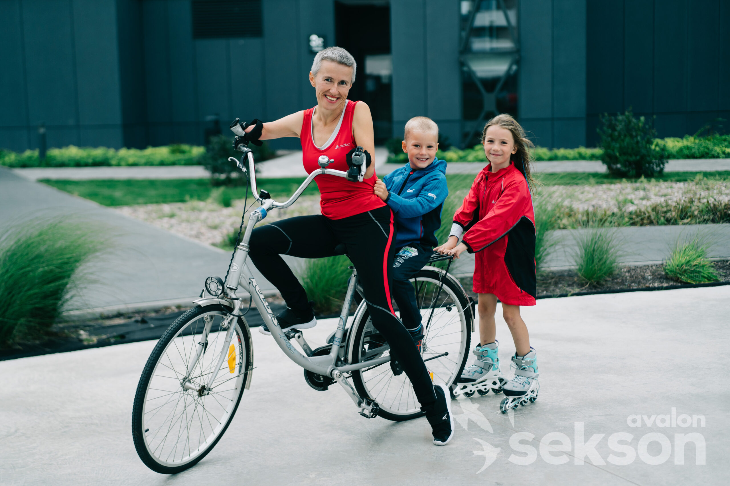 Na zdjęciu znajduje się uśmiechnięta Katarzyna Rogowiec, która jedzie rowerem. Na bagażniku siedzi mały chłopiec, a za nimi na rolkach jedzie trochę starsza dziewczynka.