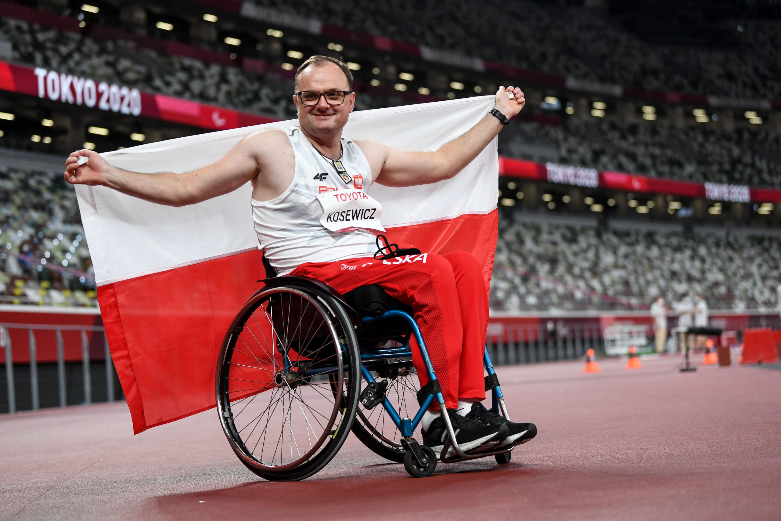 Na zdjęciu znajduje się uśmiechnięty mężczyzna na wózku (Piotr Kosewicz), który trzyma za plecami dużą flagą Polski.