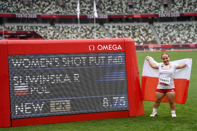 Na zdjęciu znajduje się Renata Śliwińska, trzymająca za plecami flagę Polski i stojąca na stadionie obok ekranu, na którym wyświetlają się wyniki jej rzutu.