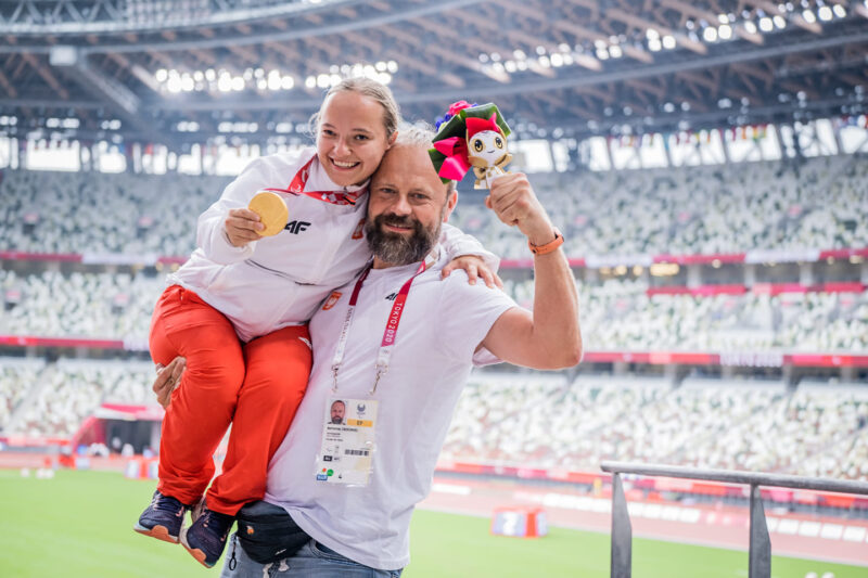 Na zdjęciu znajduje się mężczyzna, który trzyma na jednym ramieniu dziewczynę (Renatę Śliwińską), trzymającą złoty medal, wiszący na jej szyi. Oboje są uśmiechnięci, przebywają na stadionie sportowym.