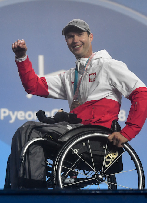 Na zdjęciu znajduje się Igor Sikorski. Siedzi na wózku, jest uśmiechnięty, podnosi rękę w górę.