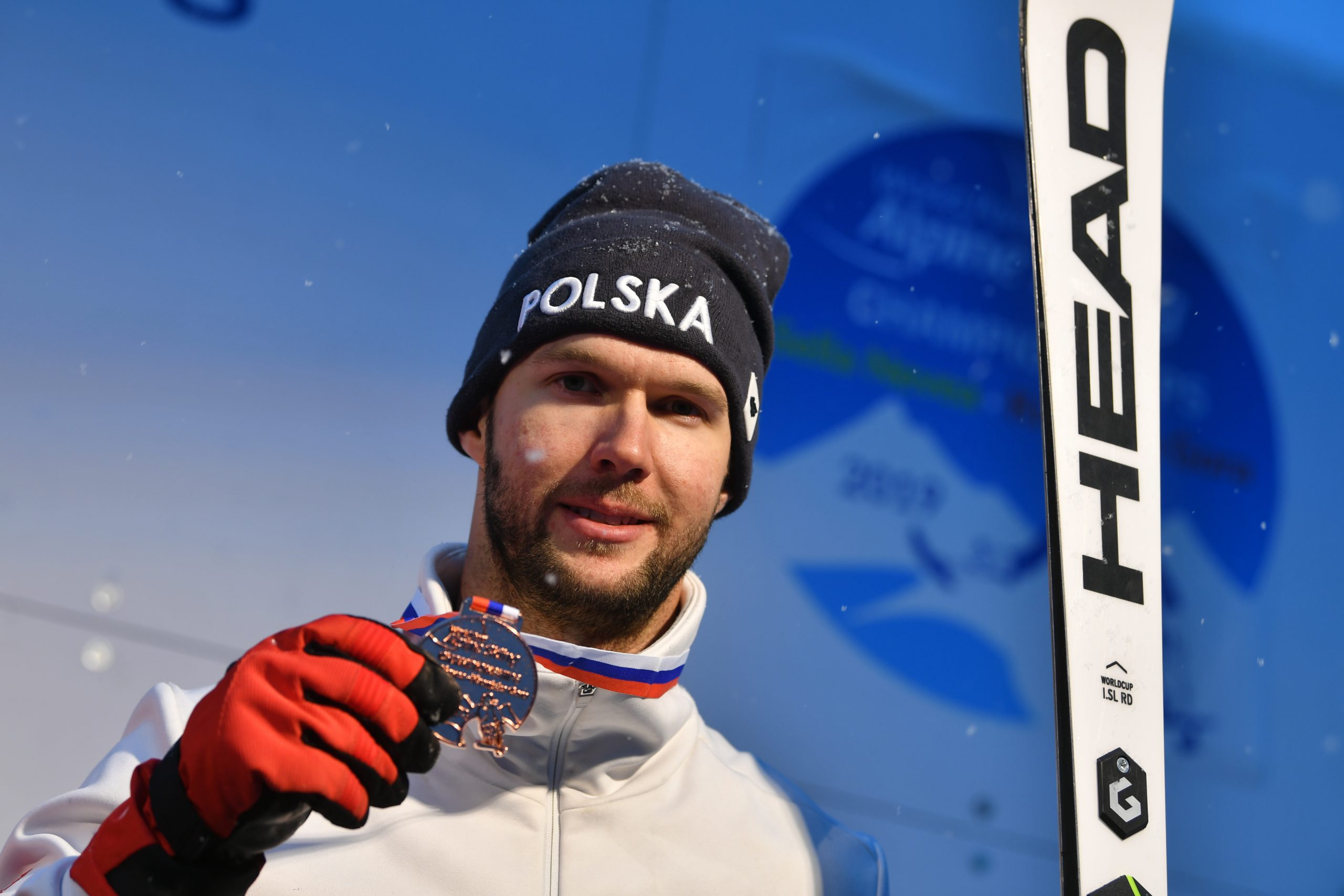 Zdjęcie portretowe mężczyzny (Igor Sikorski), który w jednej ręcze trzyma narty, a w drugiej wiszący na jego szyi medal.