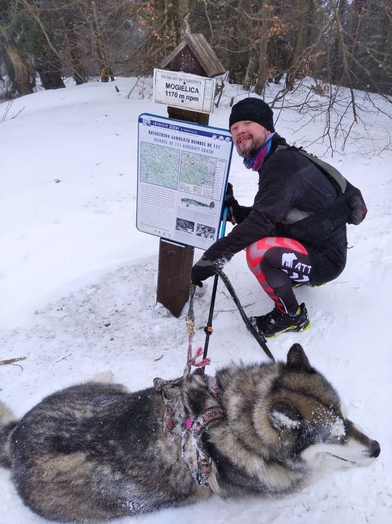 Na zdjęciu znajduje się Filip Walecki, który kuca na śniegu z psem rasy husky.