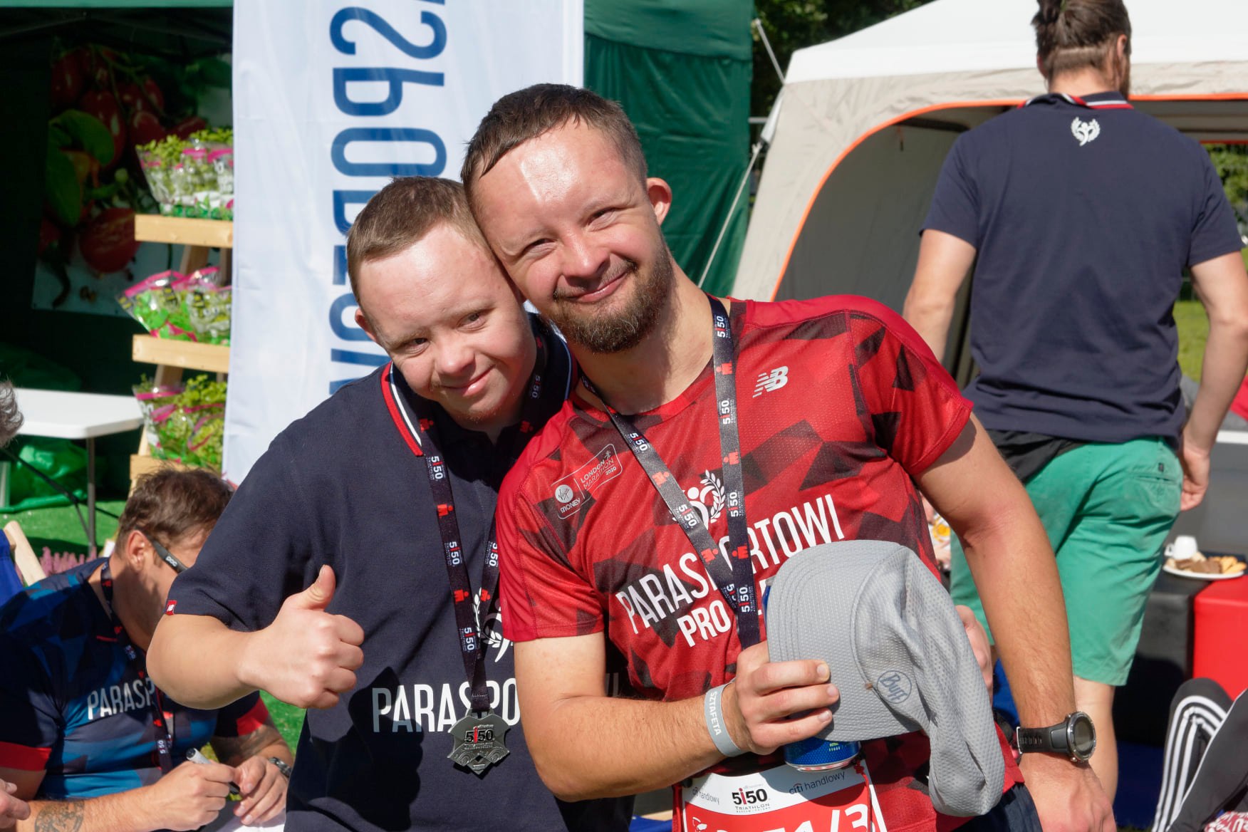 Na zdjęciu znajduje się dwóch uśmiechniętych mężczyzn z zespołem Downa w sportowych koszulkach. Jeden z nich unosi kciuk w górę.