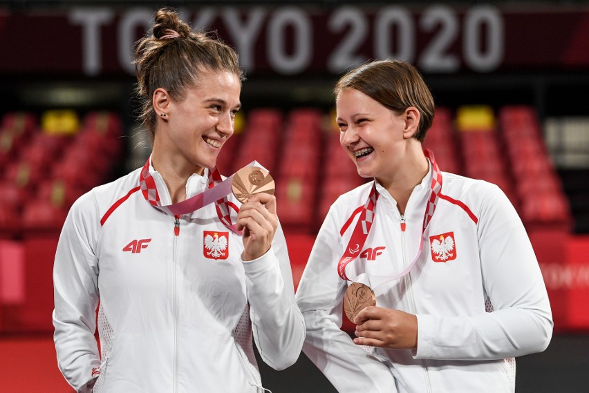 Na zdjęciu znajdują się dwie uśmiechnięte dziewczyny, które znajdują się na stadionie sportowym. Patrzą się na siebie i trzymają w rękach medale. Mają na sobie bluzy reprezentacji Polski.