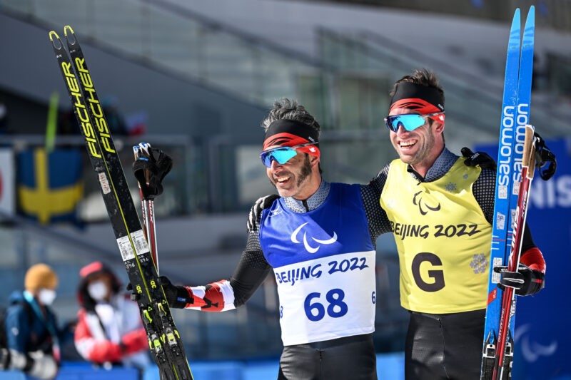 Na zdjęciu znajdują się dwaj uśmechnięci mężczyźni w strojach narciarskich. Każdy z nich trzyma w ręku parę nart, obejmują się.