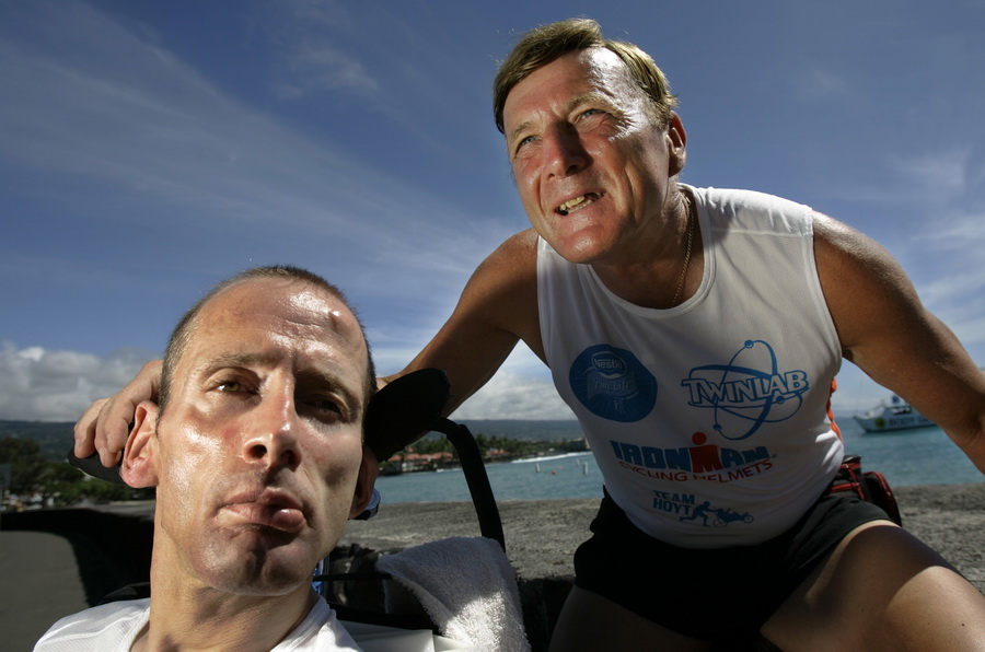 Na zdjęciu znajduje się dwóch mężczyzn na plaży: młodszy z nich jest na wózku i patrzy w obiektyw, starszy opiera się o jego wózek, uśmiecha się i patrzy w przestrzeń.