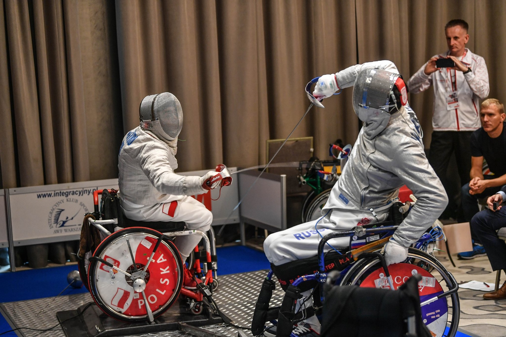 Na zdjęciu znajduje się dwóch mężczyzn na wózkach w trakcie pojedynku szermierskiego.