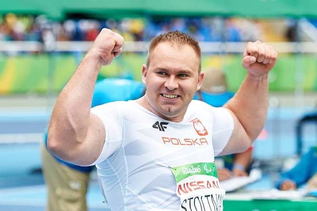 Na zdjęciu znajduje się uśmiechnięty Lech Stolatman: przebywa na stadionie sportowym, uśmiecha się i unosi ręce w górę.