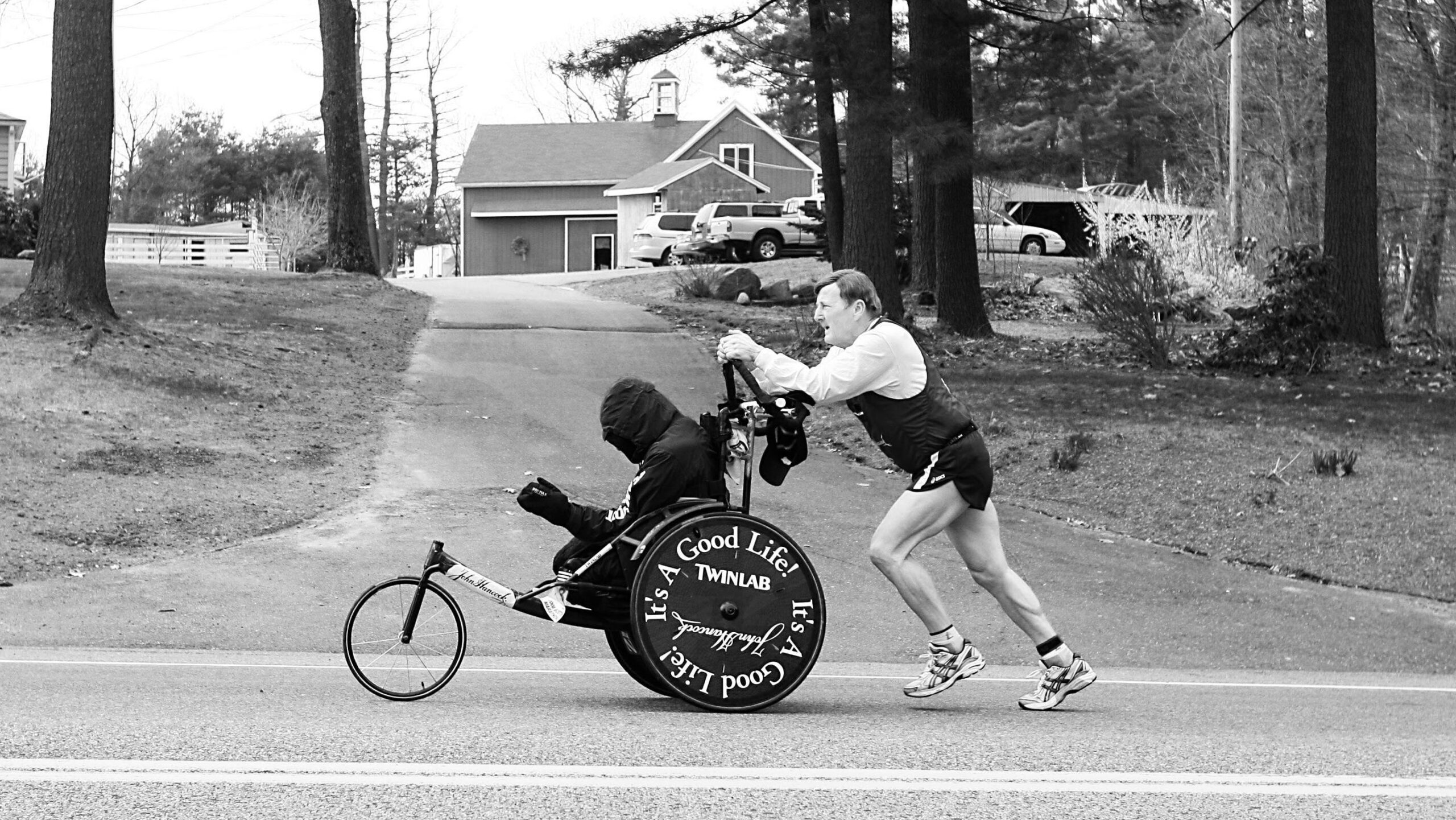 Na zdjęciu znajduje się mężczyzna w stroju biegacza, który pcha przed sobą wózek, na którym znajduje się drugi mężczyzna.