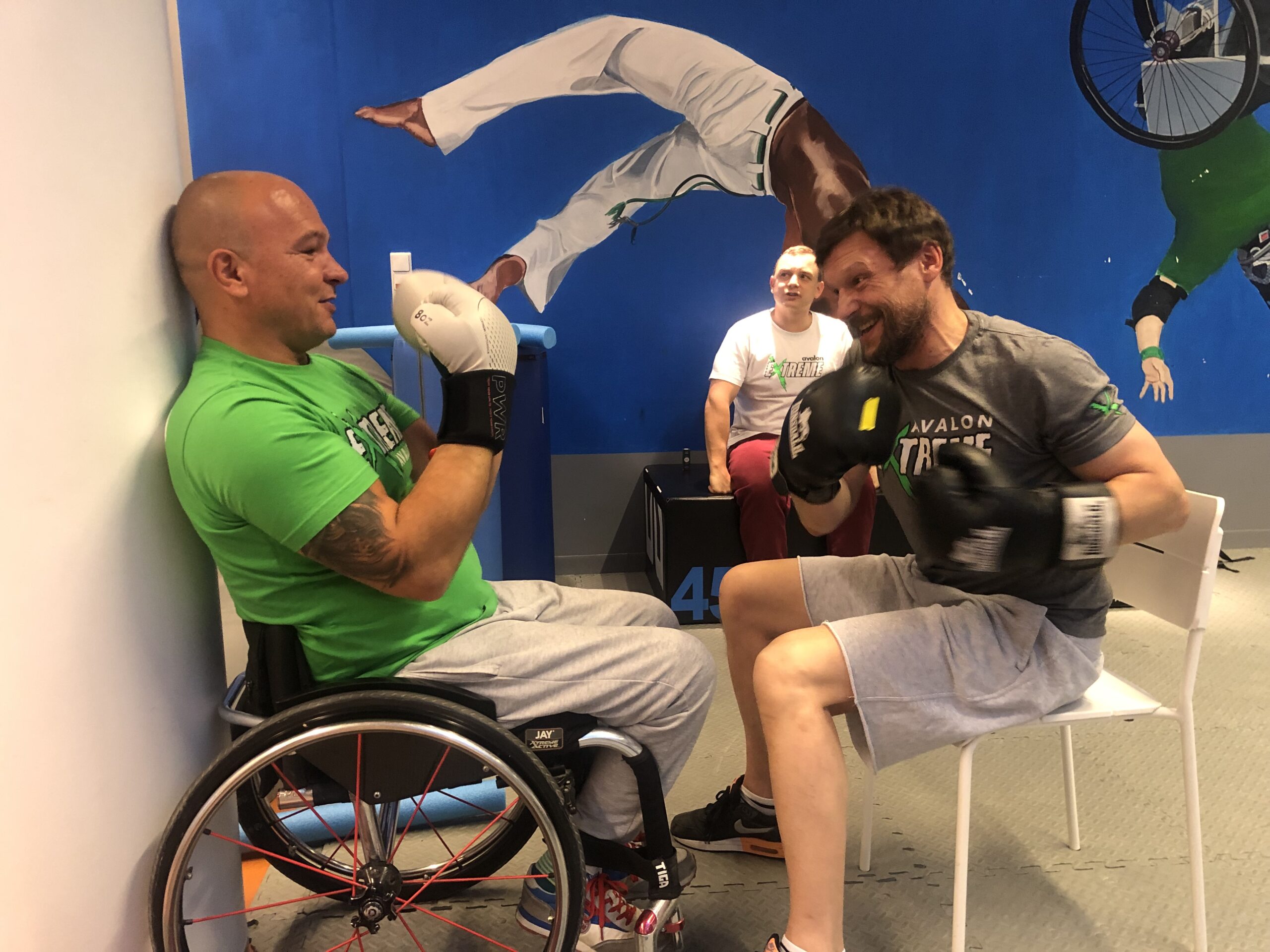Na zdjęciu znajduje się dwóch mężczyzn w rękawicach bokserskich. Jeden z nich (Arek Pilch) jest na wózku, drugi siedzi na przeciwko na krześle. Trenują boks.