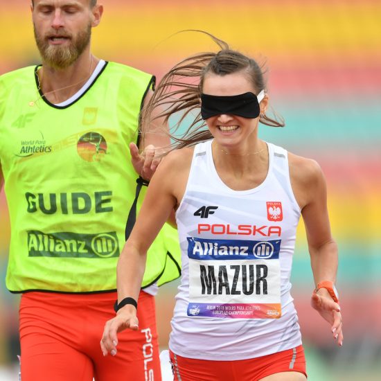 Na zdjęciu znajduje się kobieta z opaską na oczach (Joanna Mazur), która biegnie w towarzystwie swojego przewodnika.