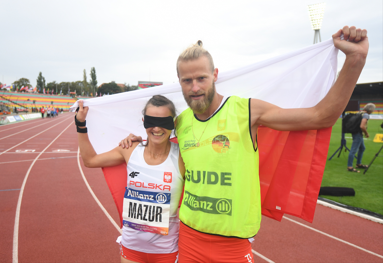 Na zdjęciu znajduje się kobieta z opaską na oczach (Joanna Mazur) i mężczyzna, który trzyma dłoń na jej ramieniu. Wspólnie trzymają dużą flagę Polski za plecami.
