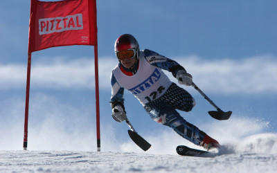 Osoba w kombinezonie narciarskim, bez lewej nogi, zjeżdża na nartach, w rękach trzyma wspierające ją kulonarty.
