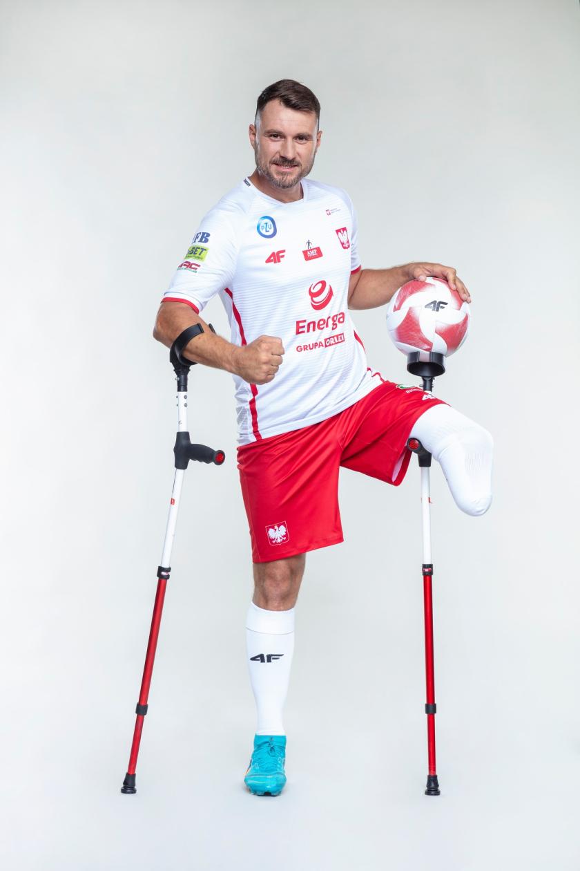 Mężczyzna w stroju reprezentacji Polski bez lewej nogi stoi z kulami i w ręku trzyma piłkę,