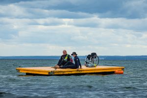Dwóch uśmiechniętych mężczyzn siedzi na pomoście na wodzie, za nimi stoi wózek inwalidzki