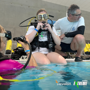 Uczestniczka nurkowania w koszulce Avalon Extreme, siedzi na brzegu basenu. Jest ubrana w pełen osprzęt do nurkowania, a obok niej znajdują się instruktorzy.