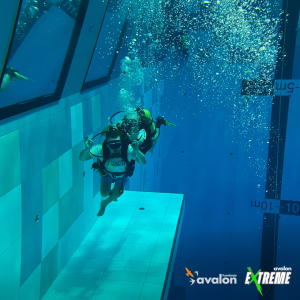 Zdjęcie uczestnika nurkowania w koszulce Avalon Extreme, zrobione pod wodą, w basenie na głębokości ok 7m. (jest to widoczne, dzięki podziałce z głębokością, zrobionej na ścianie basenu)