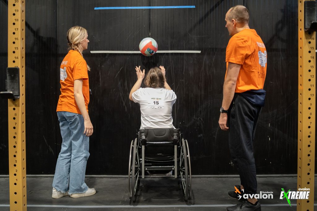 Dziewczyna na wózku odbija piłkę o ścianę, a dwójka wolontariuszy obok liczy powtórzenia.